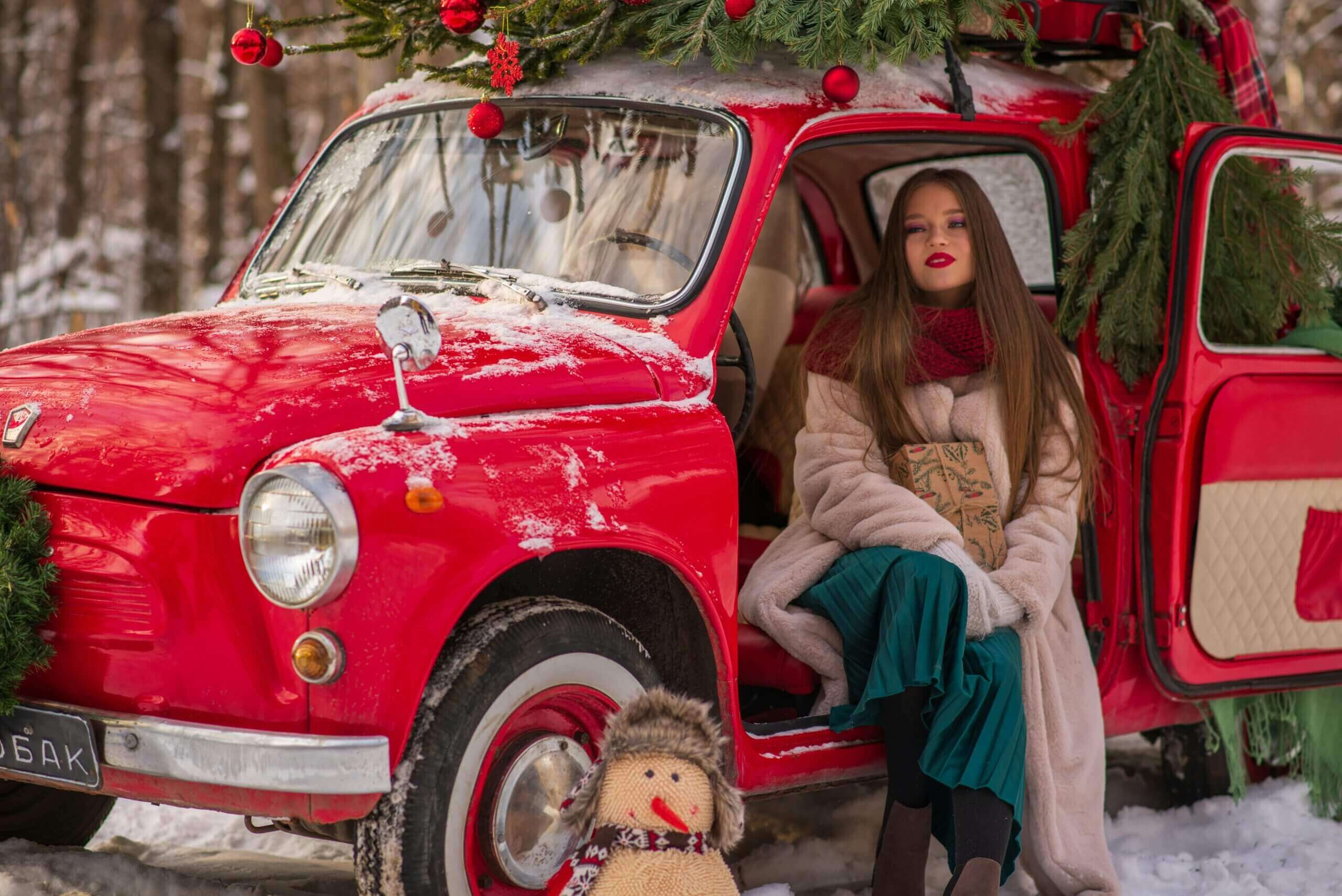 A woman in cozy winter clothes sitting by a festive red vintage car with holiday decorations.