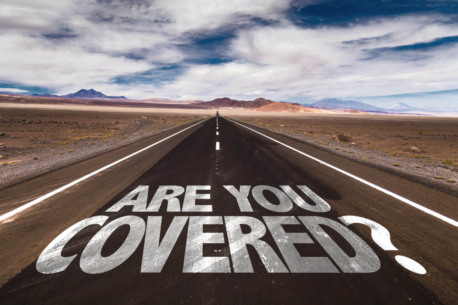A long desert road with 'ARE YOU COVERED?' painted on it under a cloudy sky.