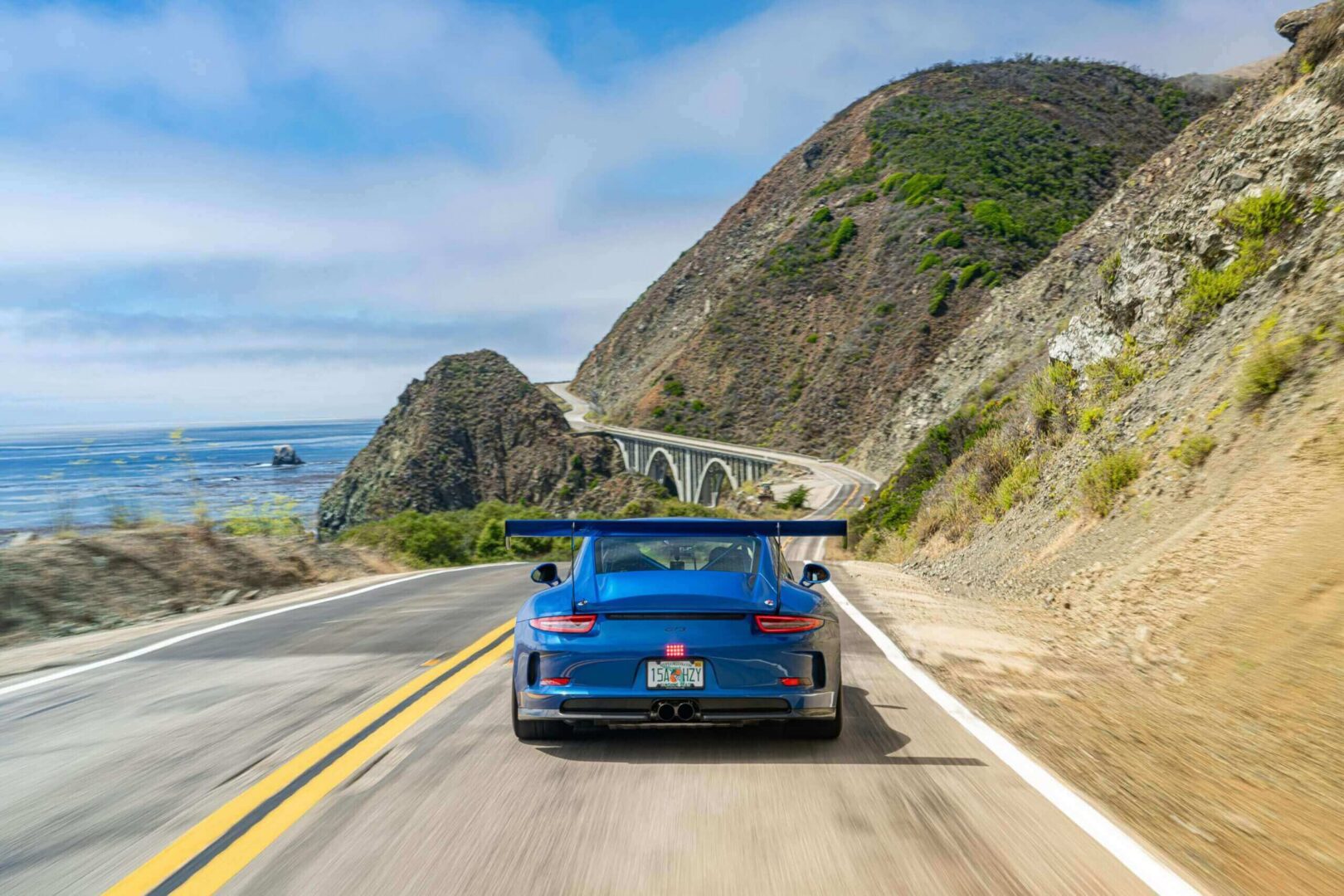 Blue sports car driving on a coastal road with bridge and hills.